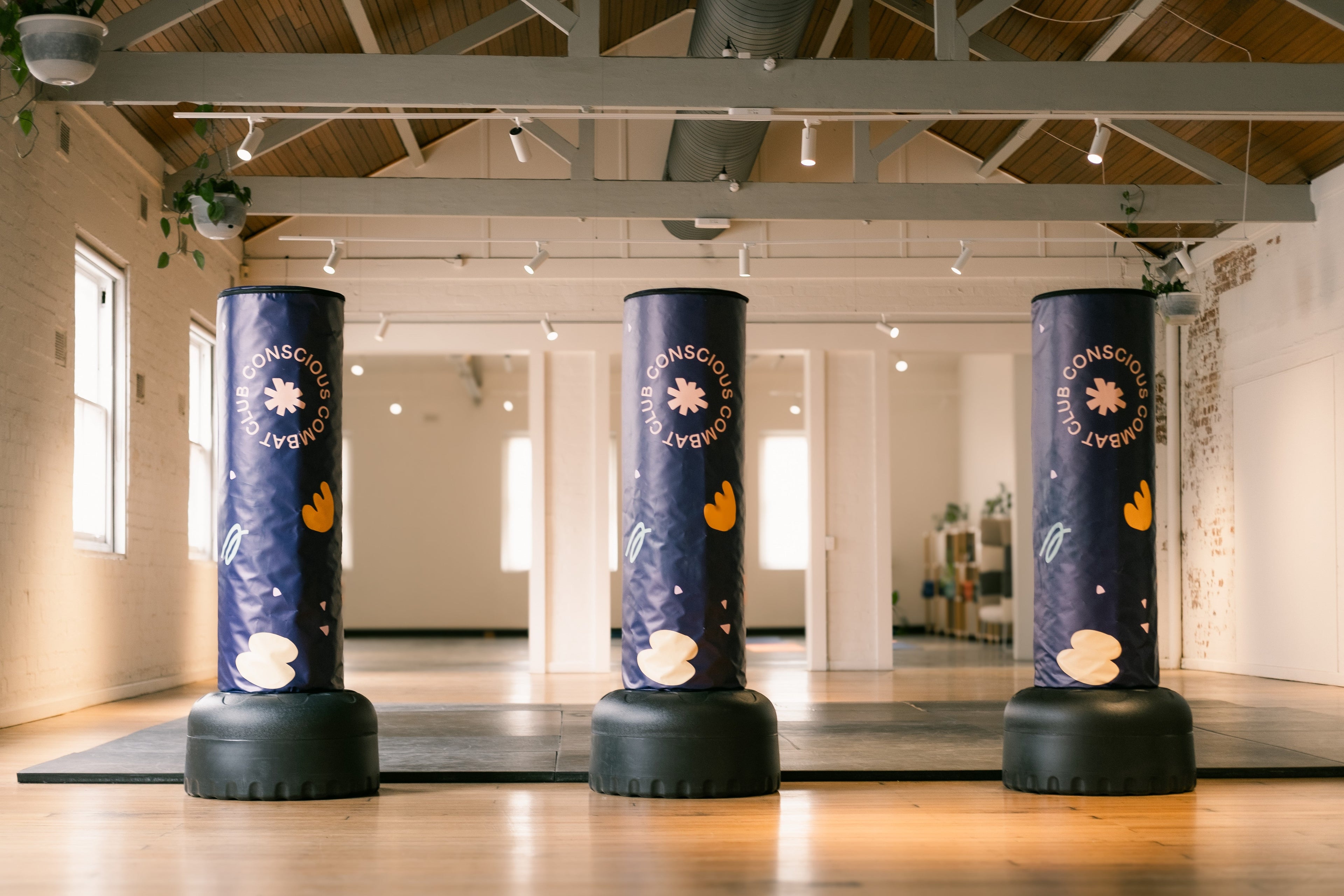Three blue punching bags with Conscious Combat Club branding in a room with wooden floors and white walls.